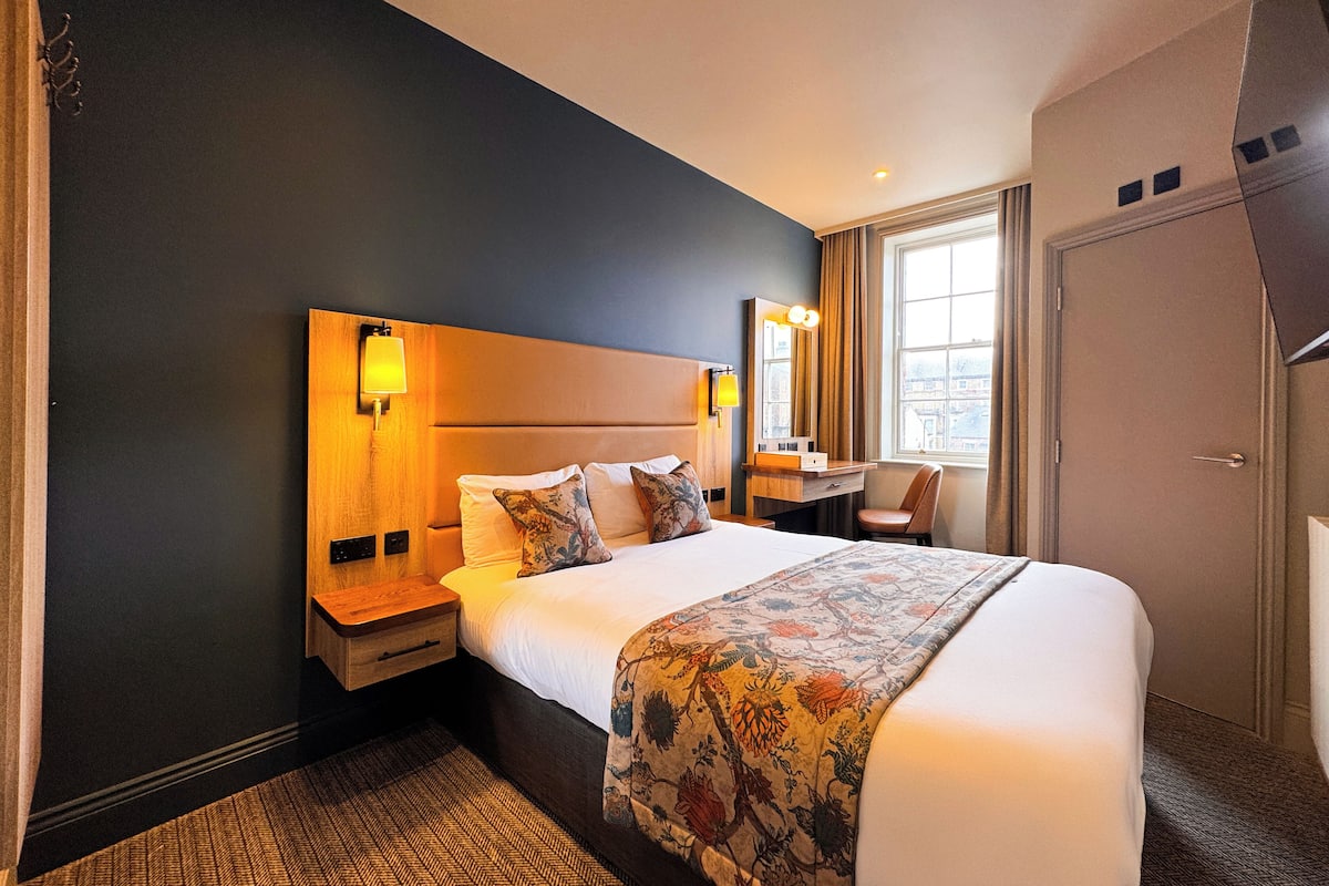 A hotel room with a large bed, a desk, a chair, and a wardrobe.