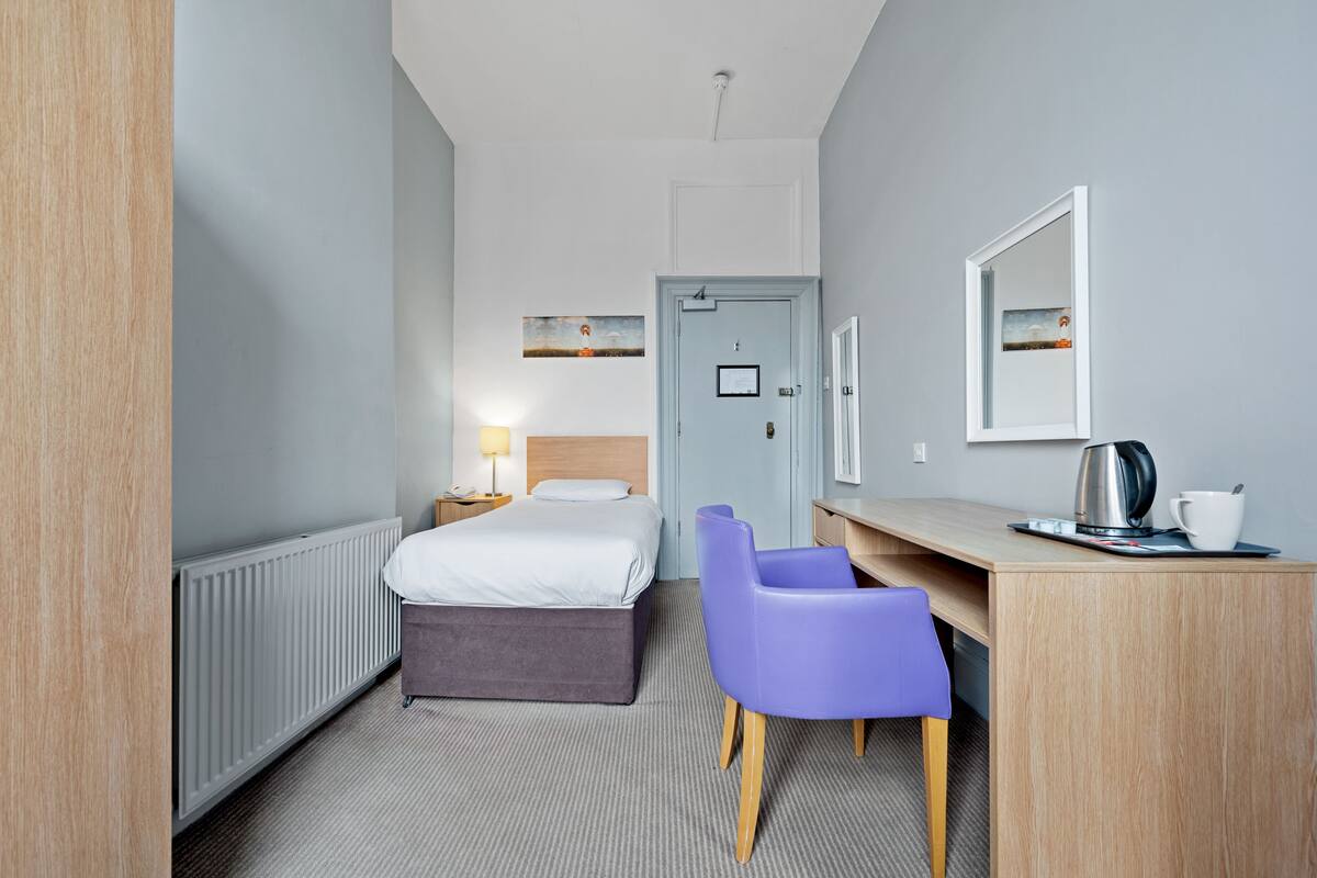 A hotel room with a bed, a desk with a chair, a radiator, and a door.