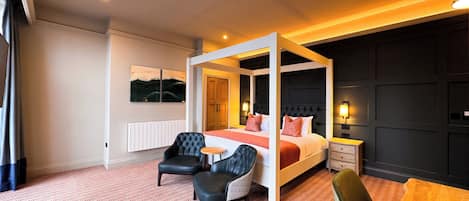 A modern hotel room with a four-poster bed, two armchairs, a small table, and a nightstand.