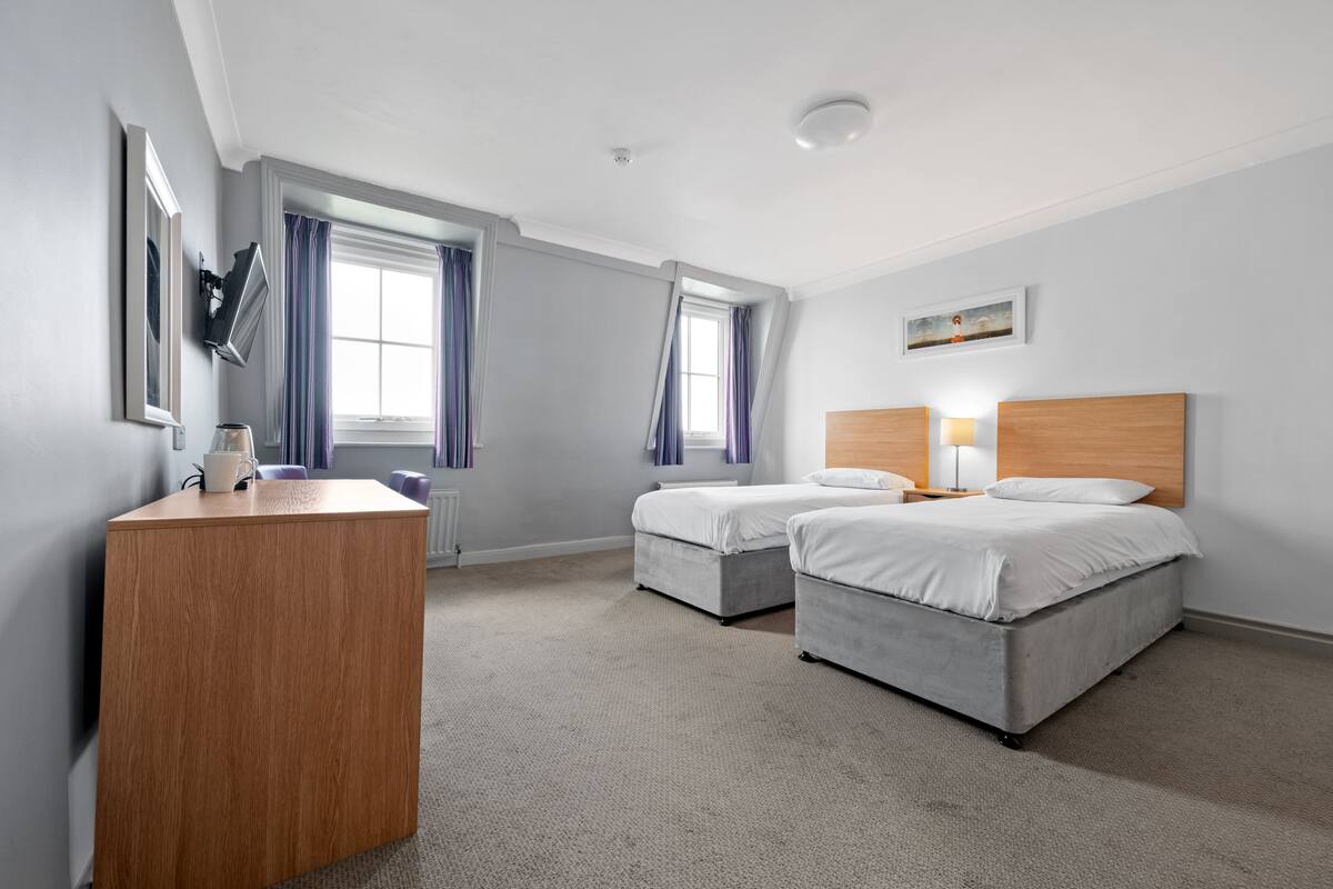 A hotel room with two beds, a wooden desk, a window with curtains, and a painting on the wall.