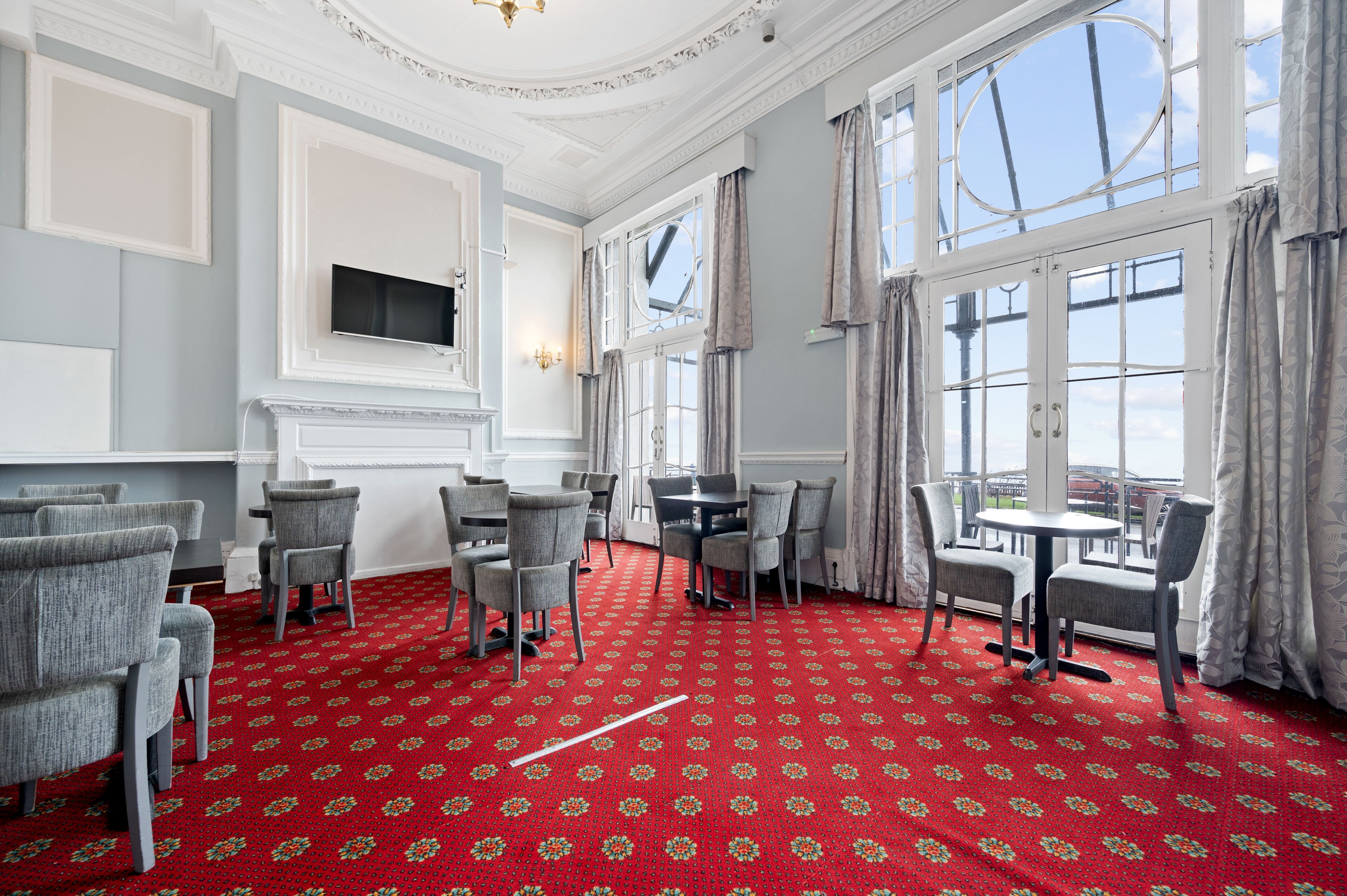 A room with a red patterned carpet, grey upholstered chairs, a round table, and large windows with curtains.