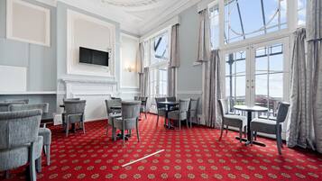 A room with a red patterned carpet, grey upholstered chairs, a round table, and large windows with curtains.