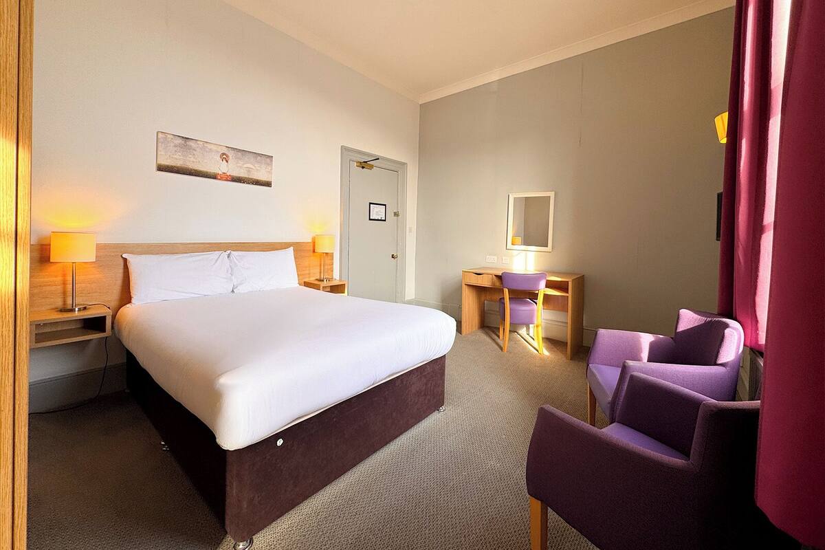 A hotel room with a bed, a desk, a chair, a lamp, and a door.