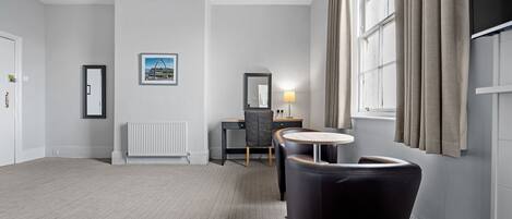 A hotel room with a large window, a desk with a chair, a small round table, and a black armchair.