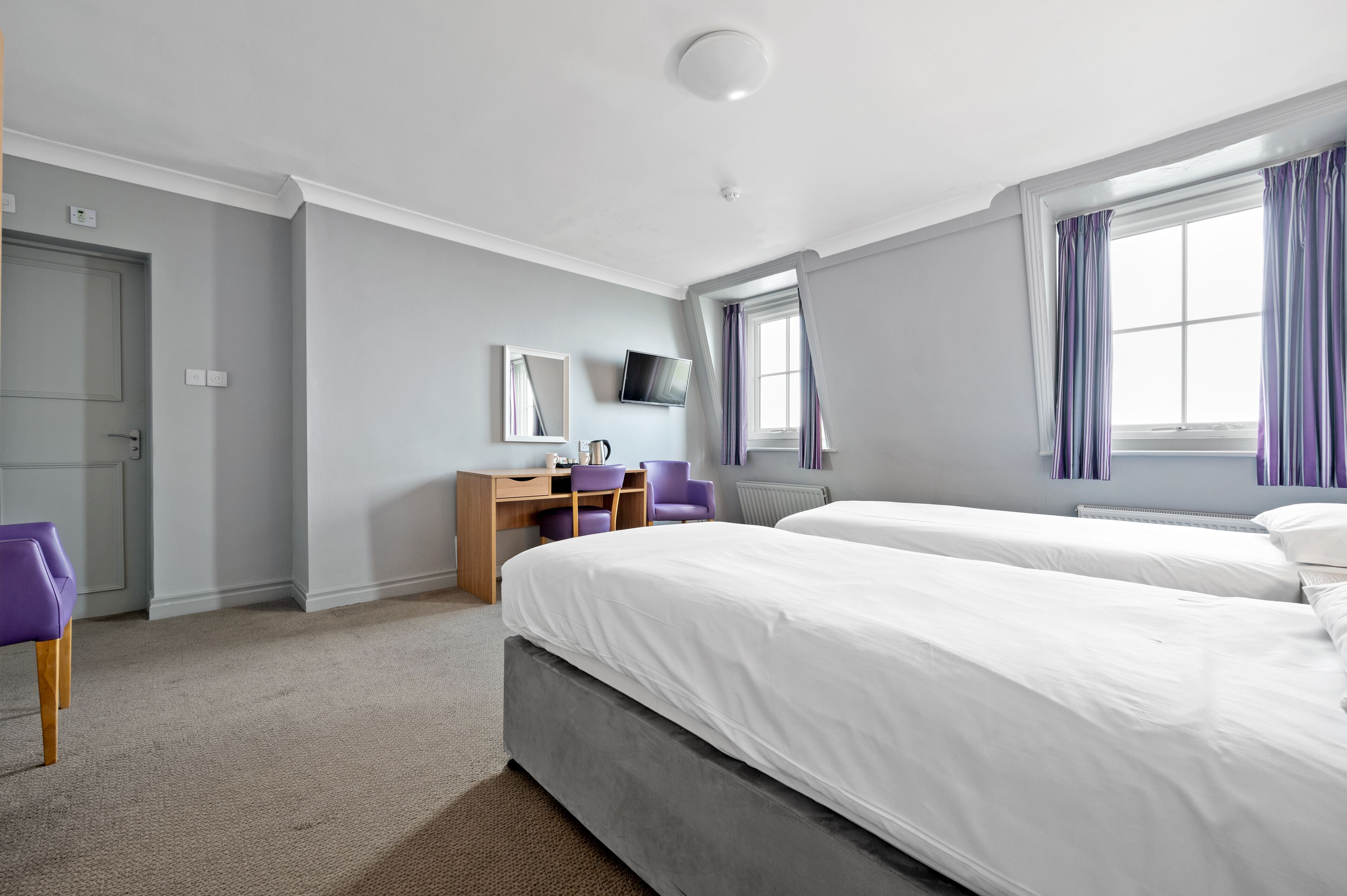 A hotel room with two beds, a desk with a chair, a TV, and a window with curtains.