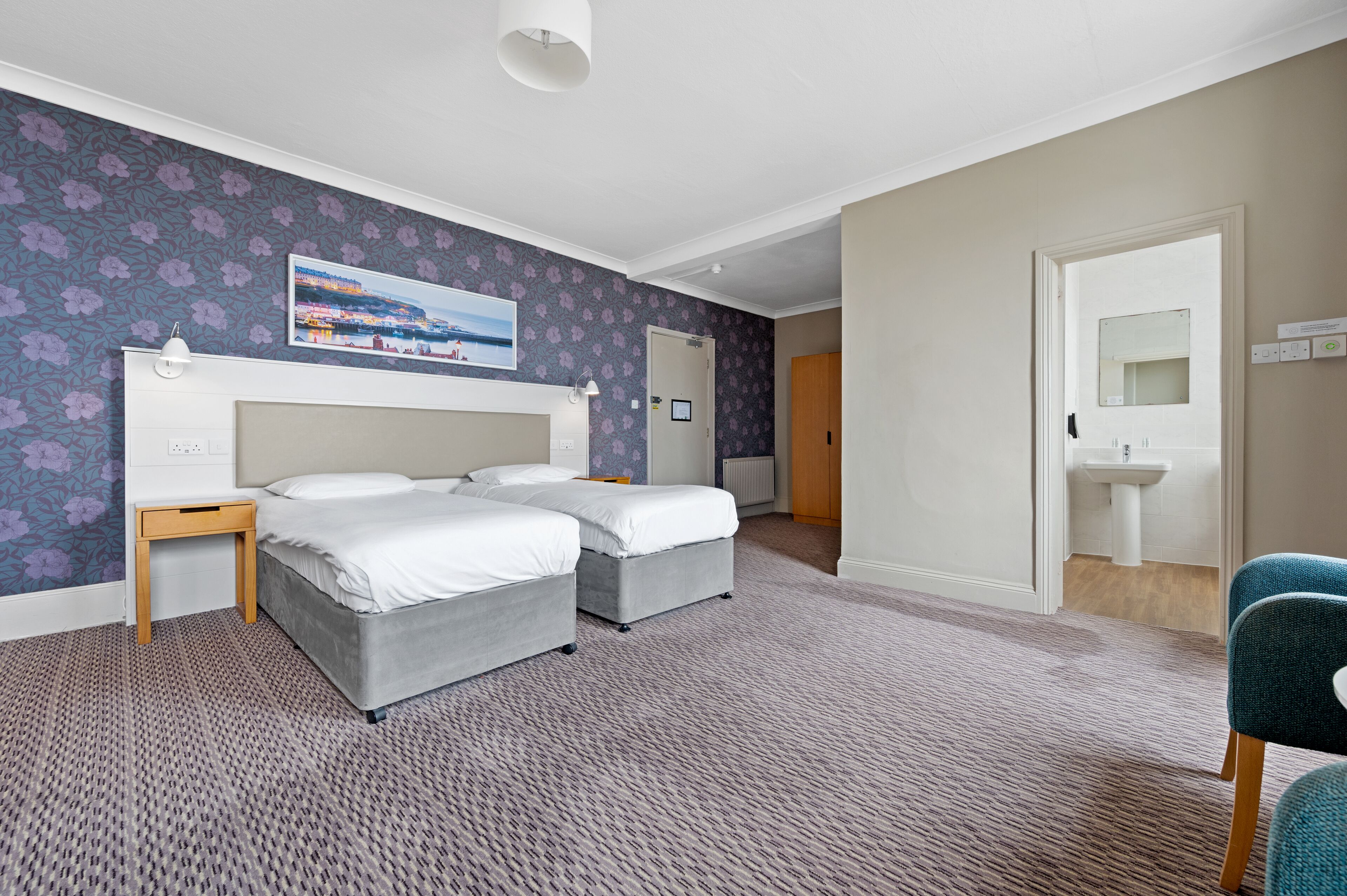 A hotel room with two beds, a small table, a chair, a painting on the wall, and a bathroom visible through an open door.