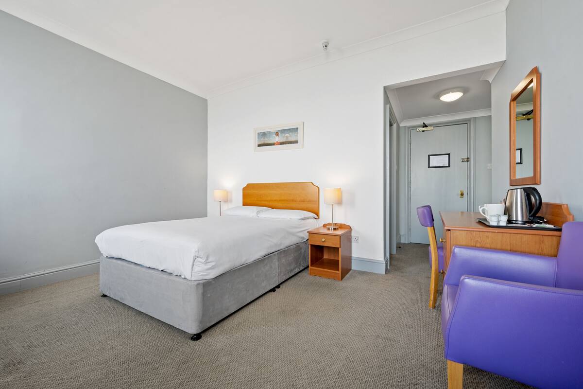 A hotel room with a bed, bedside table with lamps, a desk with a chair, and a mirror.