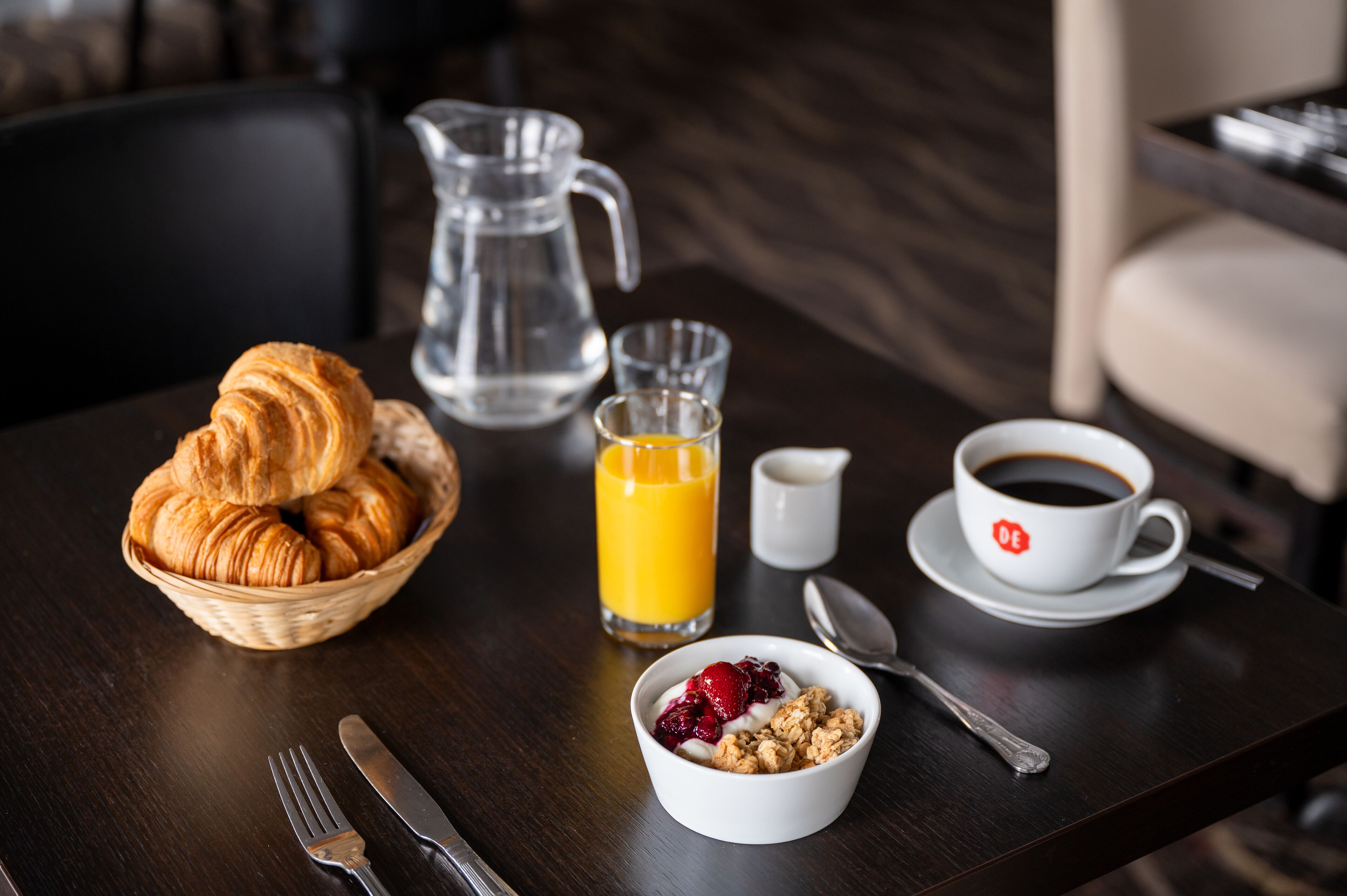 A breakfast setting with croissants, orange juice, coffee, and a bowl of granola with berries.