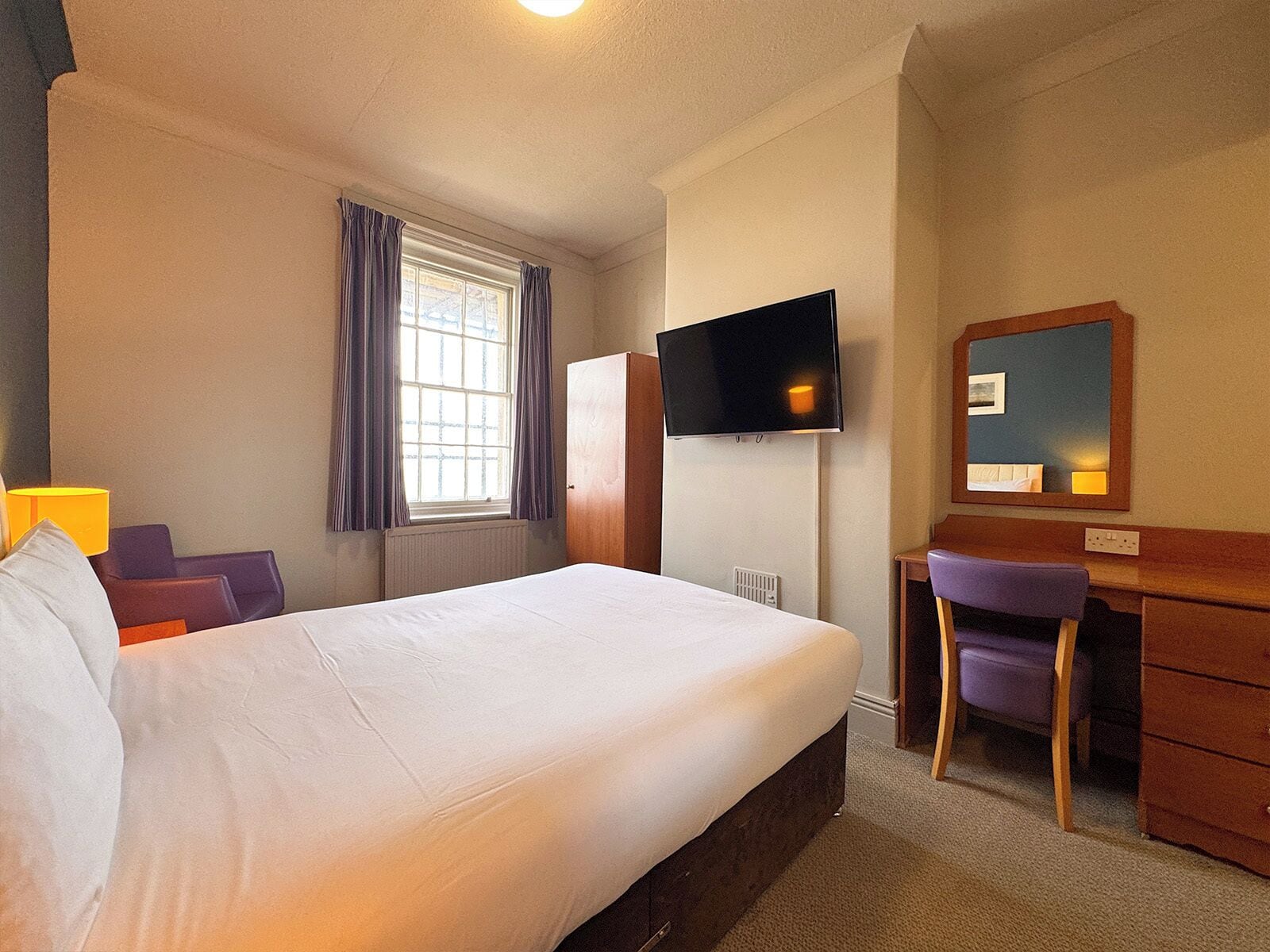 Een hotelkamer met een bed, een bureau, een televisie, een raam met gordijnen en een spiegel.