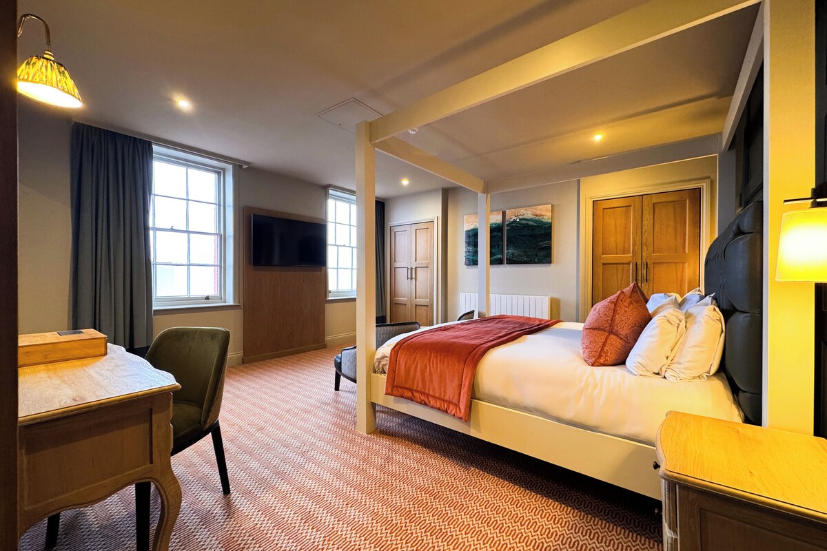 A four-poster bed with a red blanket, a desk with a chair, a television on the wall, and a window with curtains.