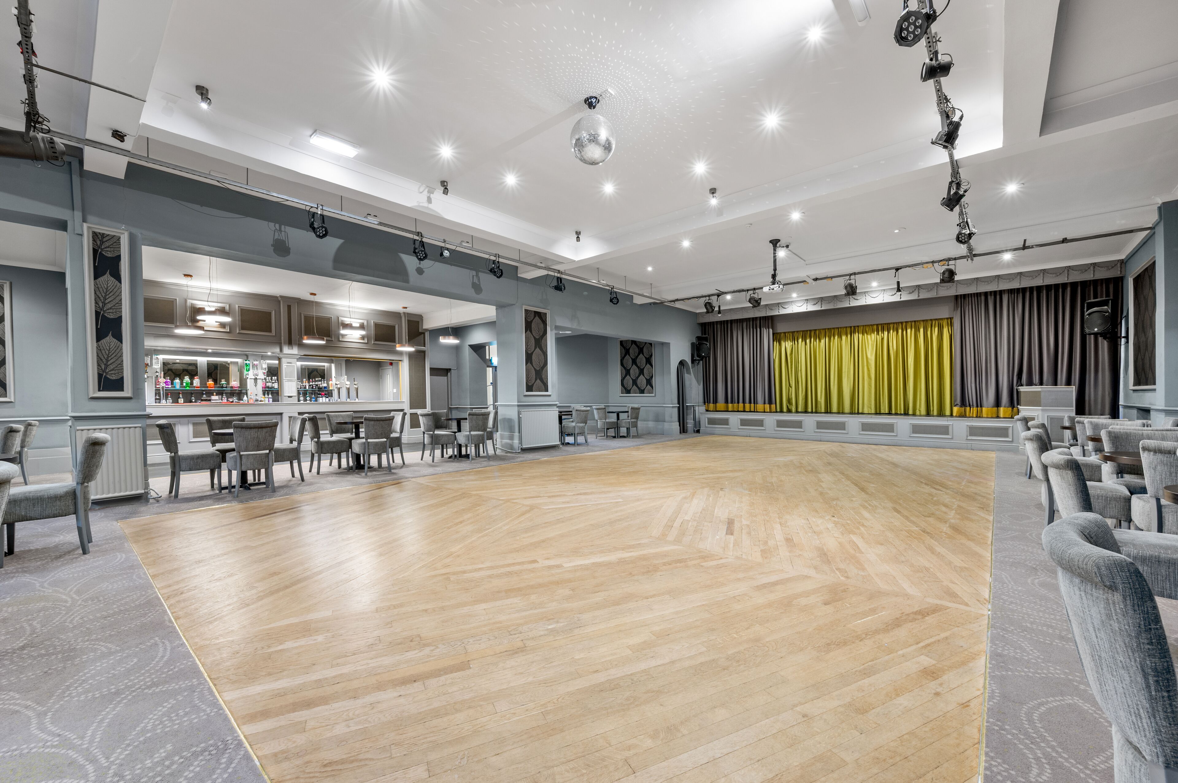 A spacious room with a wooden floor, a bar area with a counter and chairs, and a stage with curtains.