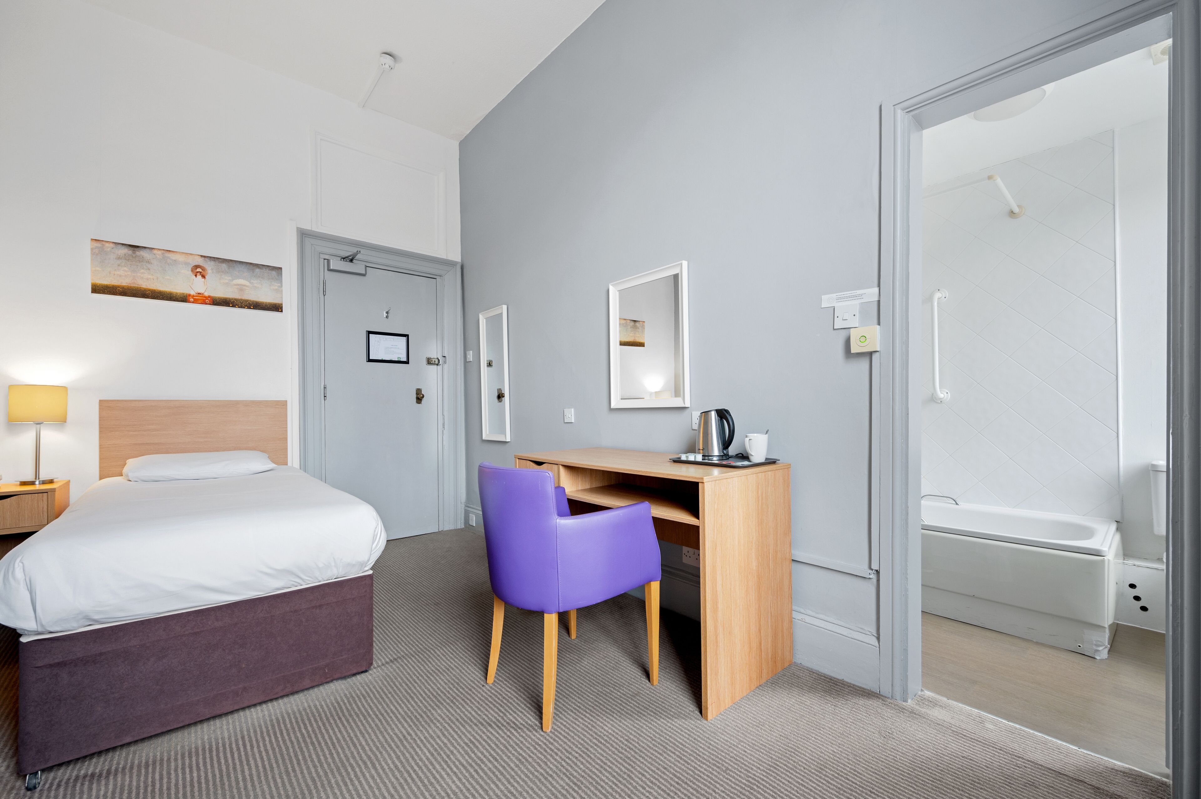 A hotel room with a bed, a desk with a chair, a shower, and a painting on the wall.