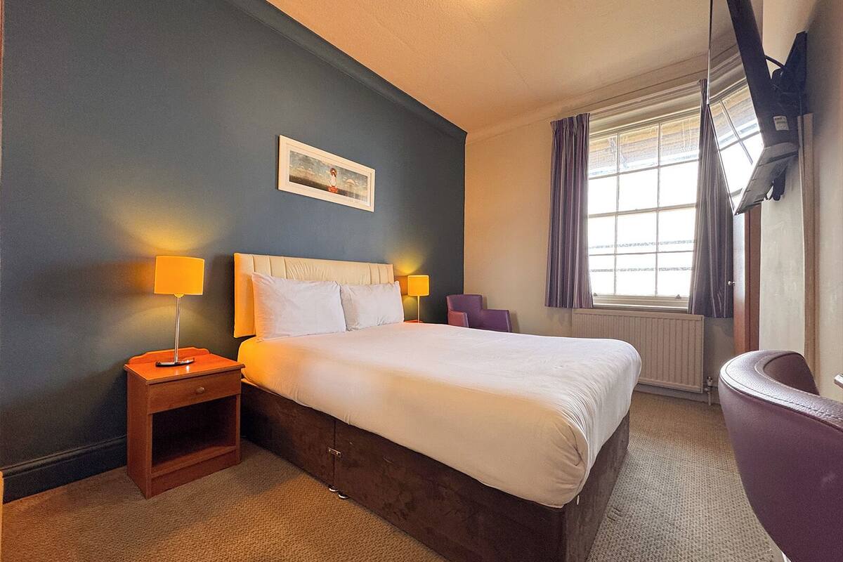 A hotel room with a bed, two bedside lamps, a framed picture on the wall, a window with curtains, and a small seating area with a chair.