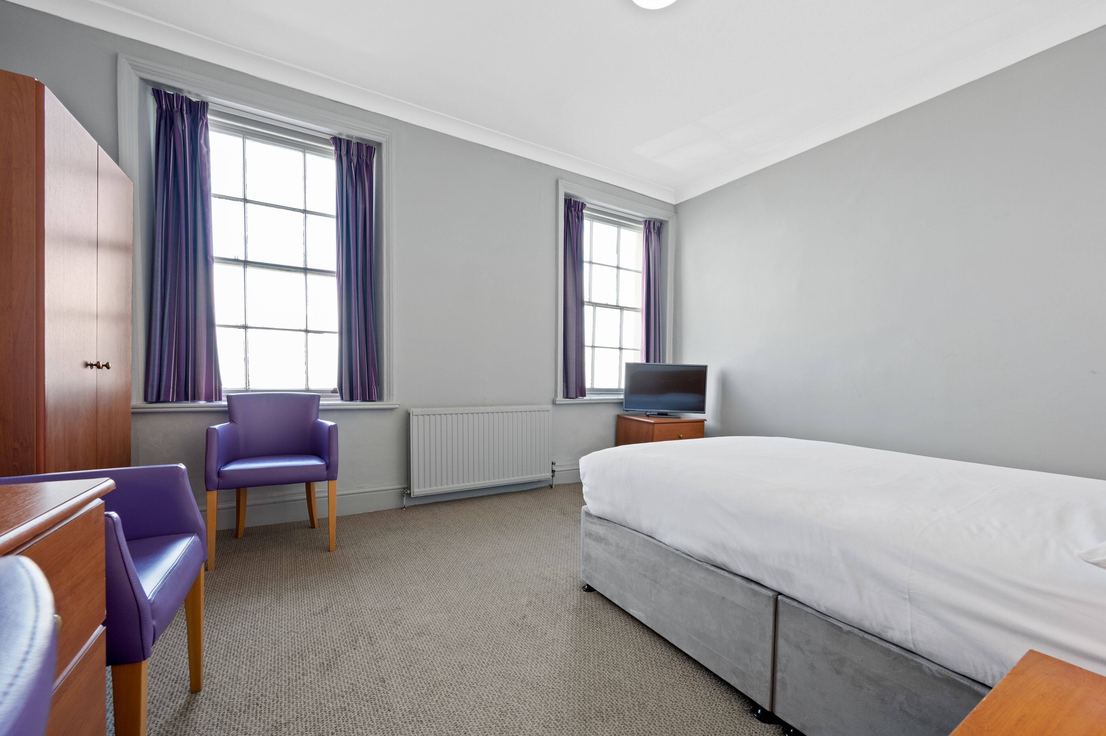 A hotel room with a bed, a TV, a chair, and a wardrobe.