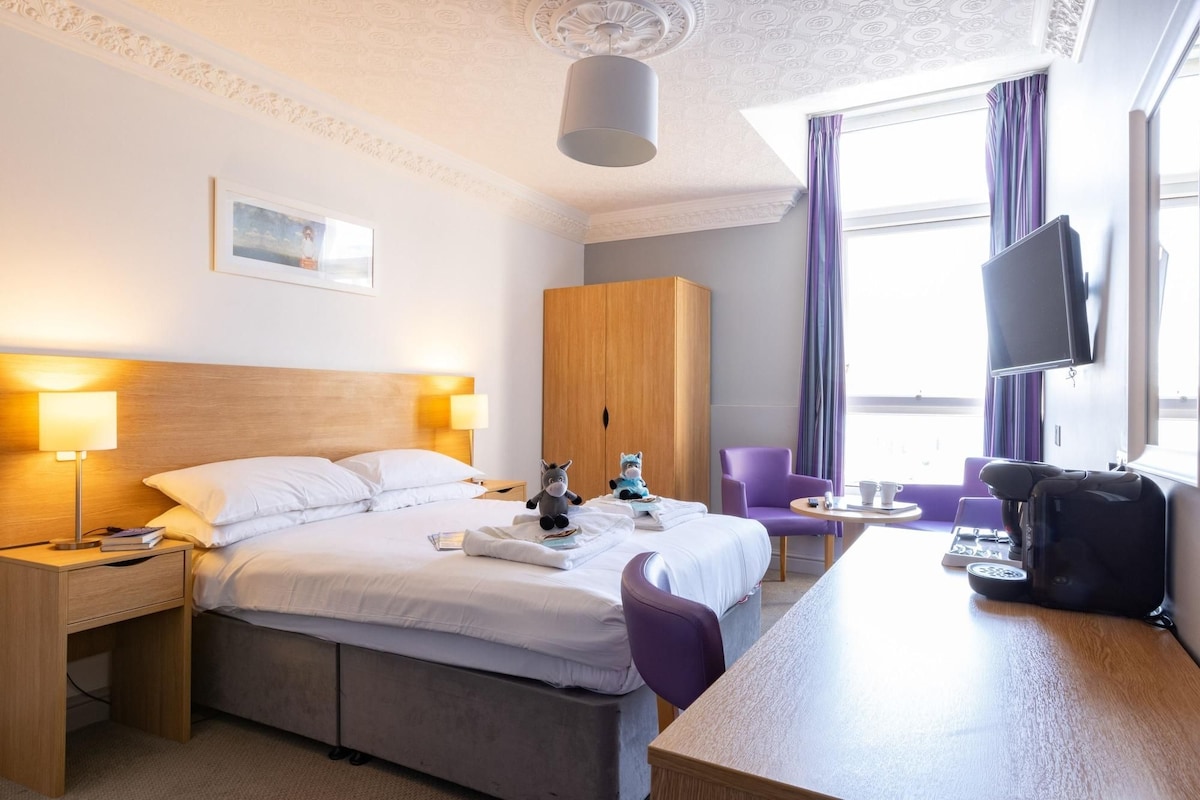 Habitación de hotel con cama, mesitas de noche, escritorio, silla, armario, televisor y ventana con cortinas.