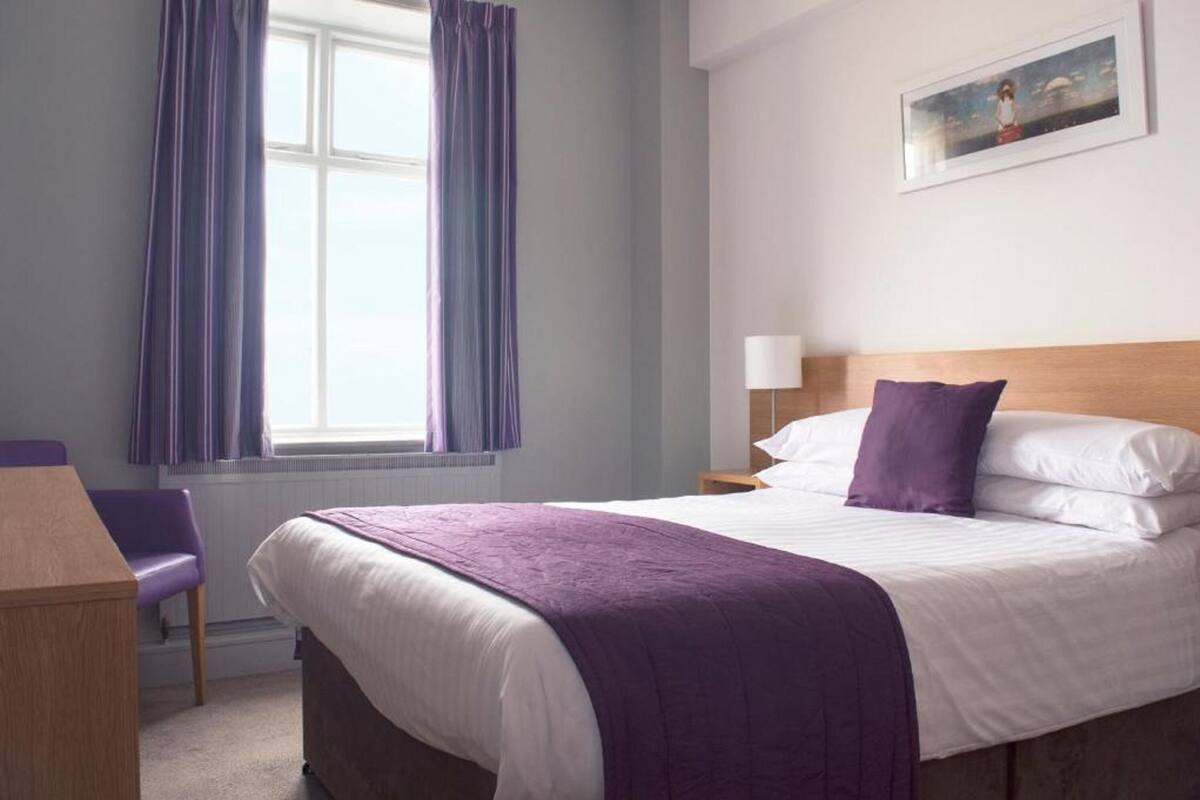 A hotel room with a bed, a chair, a nightstand, a lamp, and a window with curtains.