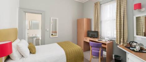 Quarto de hotel com cama, escrivaninha, cadeira, computador e banheiro pequeno.