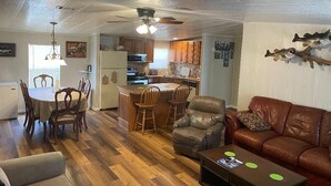 Ein geräumiges Wohnzimmer mit einer braunen Ledersofa, einem Essbereich mit Holzsesseln, einer Küche mit Holzausstattung und einem Deckenventilator.