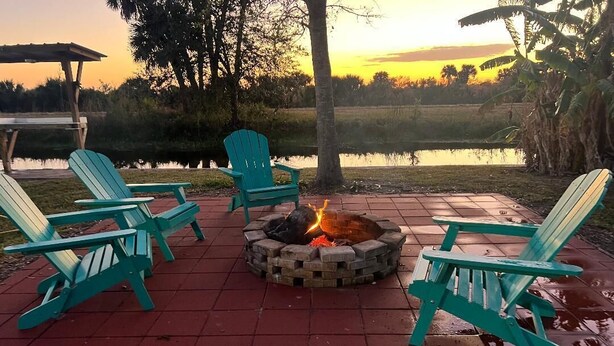 Ein Feuerkorb mit vier Adirondack-Stühlen darum herum, auf einer Ziegelterrasse mit Blick auf einen See und den Sonnenuntergang.