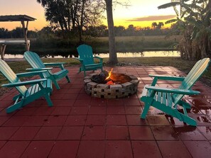 A fire pit with four Adirondack chairs around it, set on a brick patio with a view of a lake and sunset. - Comfortable Okeechobee Home Minutes From Scott Driver, Okee-Tantie Boat Ramp (Okeechobee)
