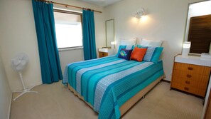 A bedroom with a bed, a bedside table, a mirror, a fan, and a window with curtains. - Ashwood | Ocean Views Opposite Kings Beach (KINGS BEACH)