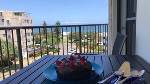 A balcony with a table holding a chocolate cake with strawberries, overlooking a residential area with buildings and a clear sky.
