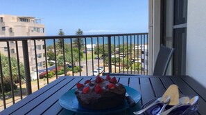 Un balcón con una mesa que sostiene un pastel de chocolate con fresas, con vistas a una zona residencial con edificios y un cielo despejado.