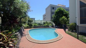 A swimming pool with a curved edge, surrounded by a red-tiled deck, with a building and greenery in the background. - Ashwood | Ocean Views Opposite Kings Beach (KINGS BEACH)