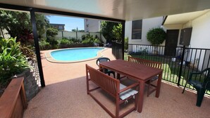 An outdoor patio with a pool, wooden furniture, and a fenced garden area.