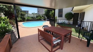 Un patio exterior con piscina, muebles de madera y un jardín cercado.