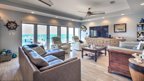 A spacious living room with a large sofa, two armchairs, a wooden coffee table, and a television. There are ceiling fans, a large window with a view of the ocean, and a bookshelf.