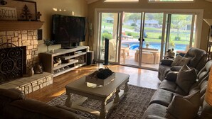 A spacious living room with a stone fireplace, a flat-screen TV, and a large sliding glass door leading to a pool area. - On the Golf Course Home w amazing views!  (San Jose)