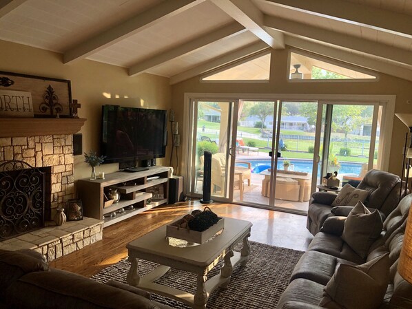 A spacious living room with a stone fireplace, a flat-screen TV, and a large sliding glass door leading to a pool area.