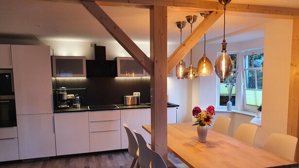 A modern kitchen with white cabinets, a black countertop, and stainless steel appliances. There is a dining area with a wooden table and white chairs, and a vase with flowers on the table. - Gute Laune Hof Klingenthal (Klingenthal)