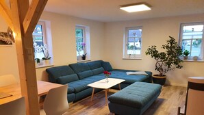 A modern living room with a blue sofa, a white coffee table, and a dining table with chairs. There are windows with plants and a view of the outside. - Gute Laune Hof Klingenthal (Klingenthal)