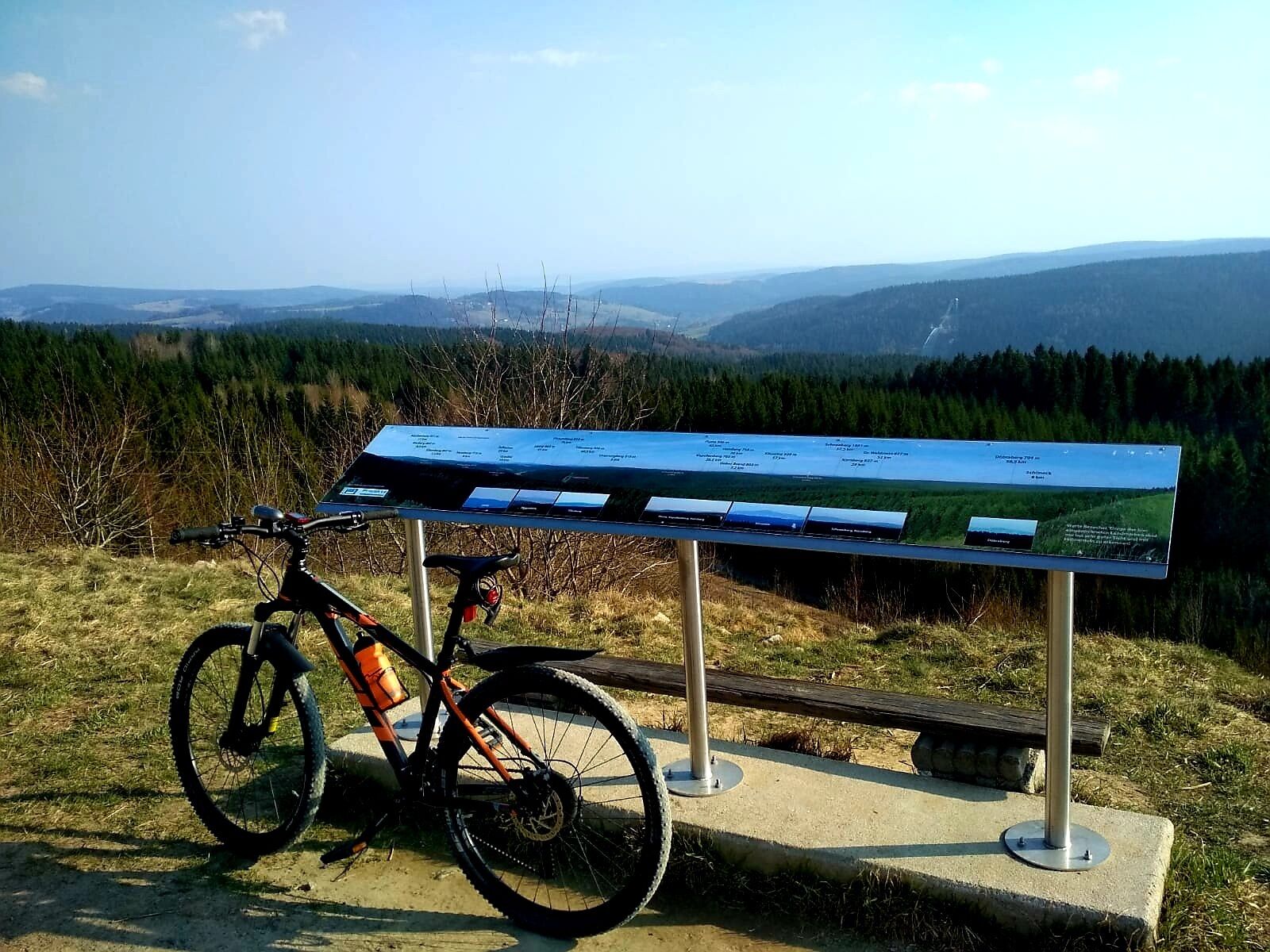 Una bicicleta de montaña estacionada junto a un panel informativo metálico, con una vista panorámica de un valle boscoso y montañas distantes.