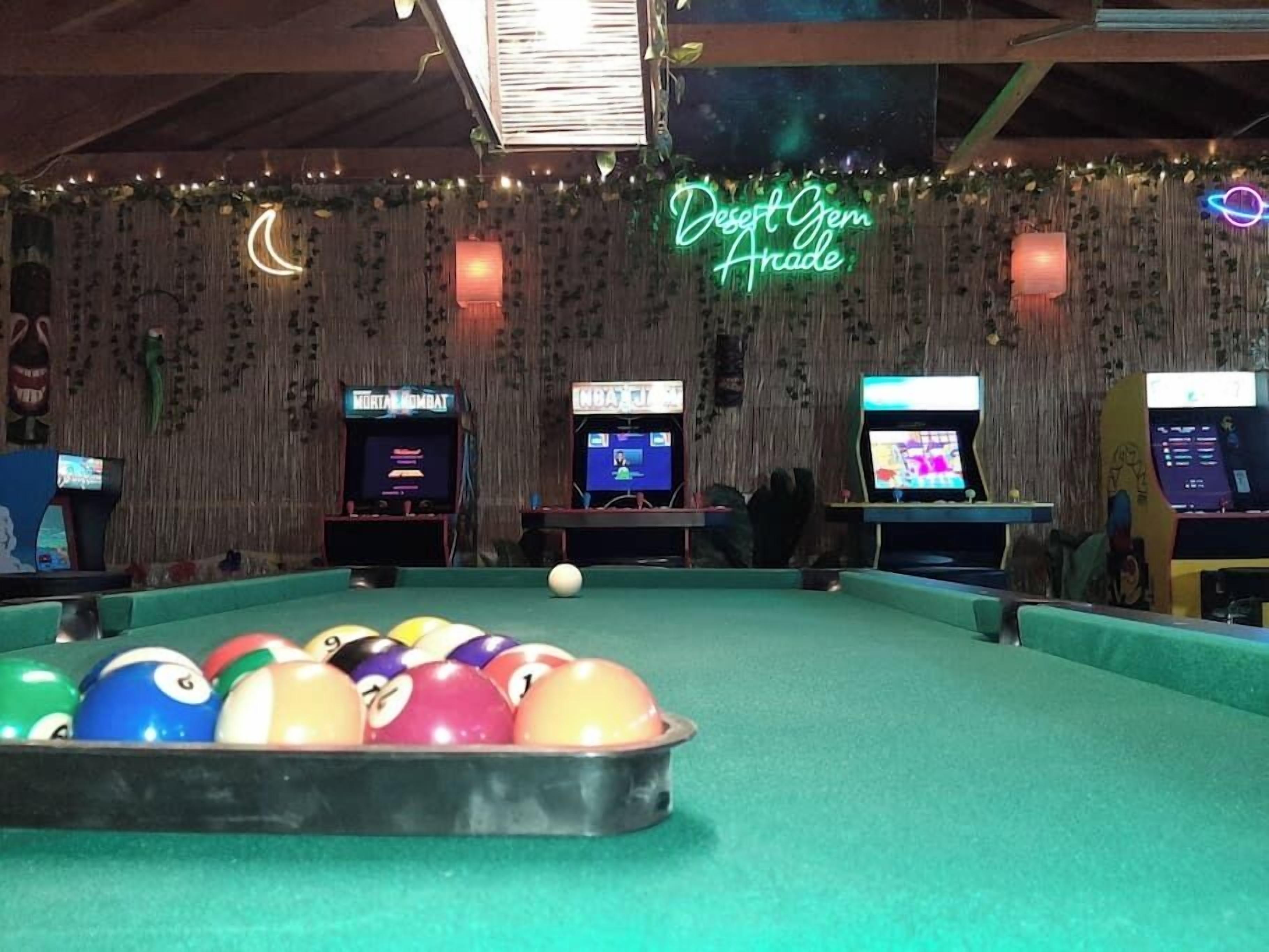 A pool table with colorful balls, arcade machines, and a neon sign reading "Desert Gem Arcade".