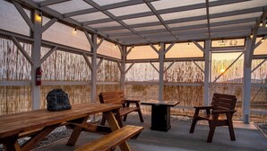 Een overdekt terras met houten banken en picknicktafels, een brandblusser en uitzicht op de zonsondergang door een versierd hek.