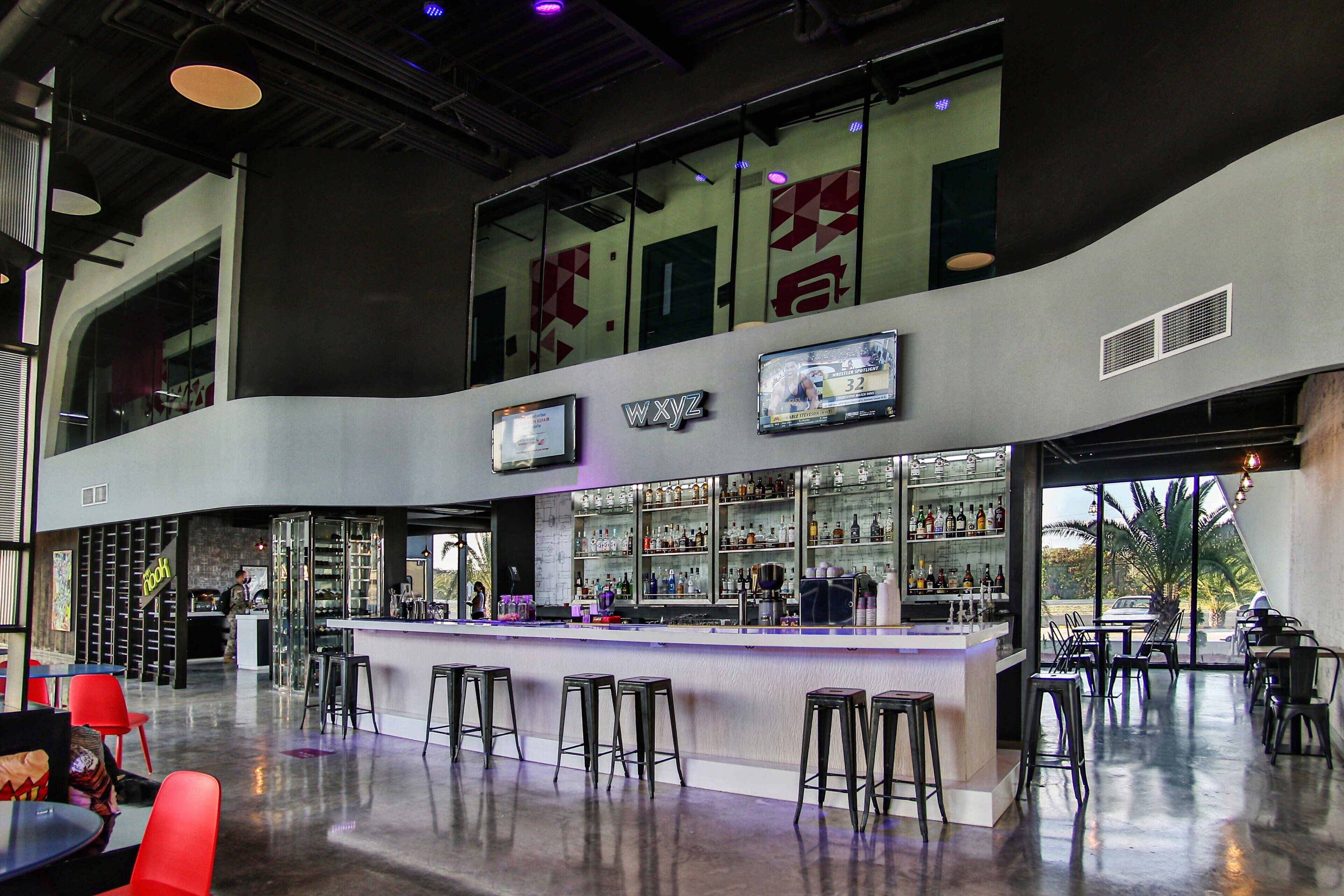 Un área de bar moderna con techo alto, barra, varios taburetes y un amplio ventanal con vistas al exterior.