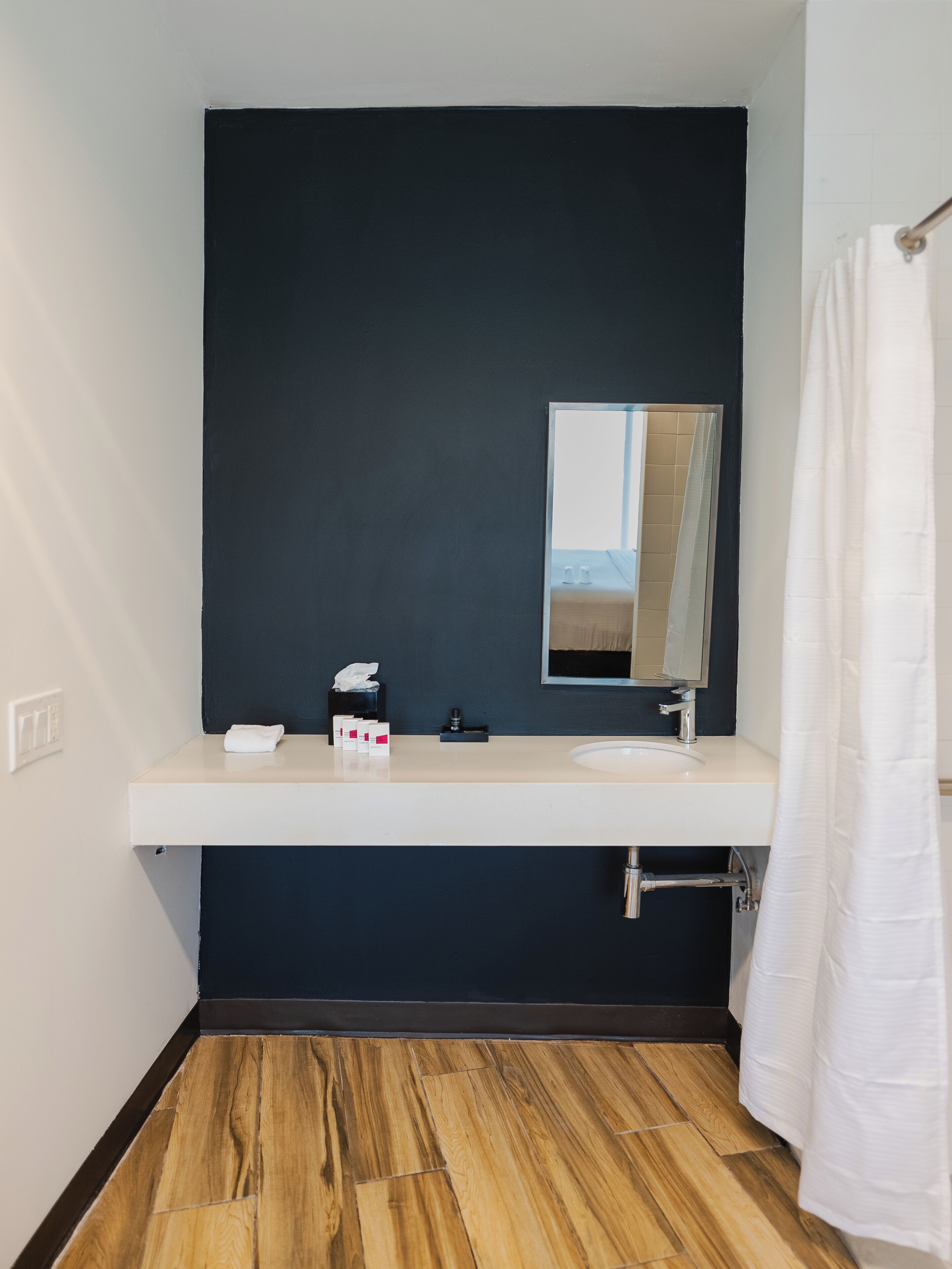 A bathroom with a dark wall, a white sink, a mirror, and a wooden floor.