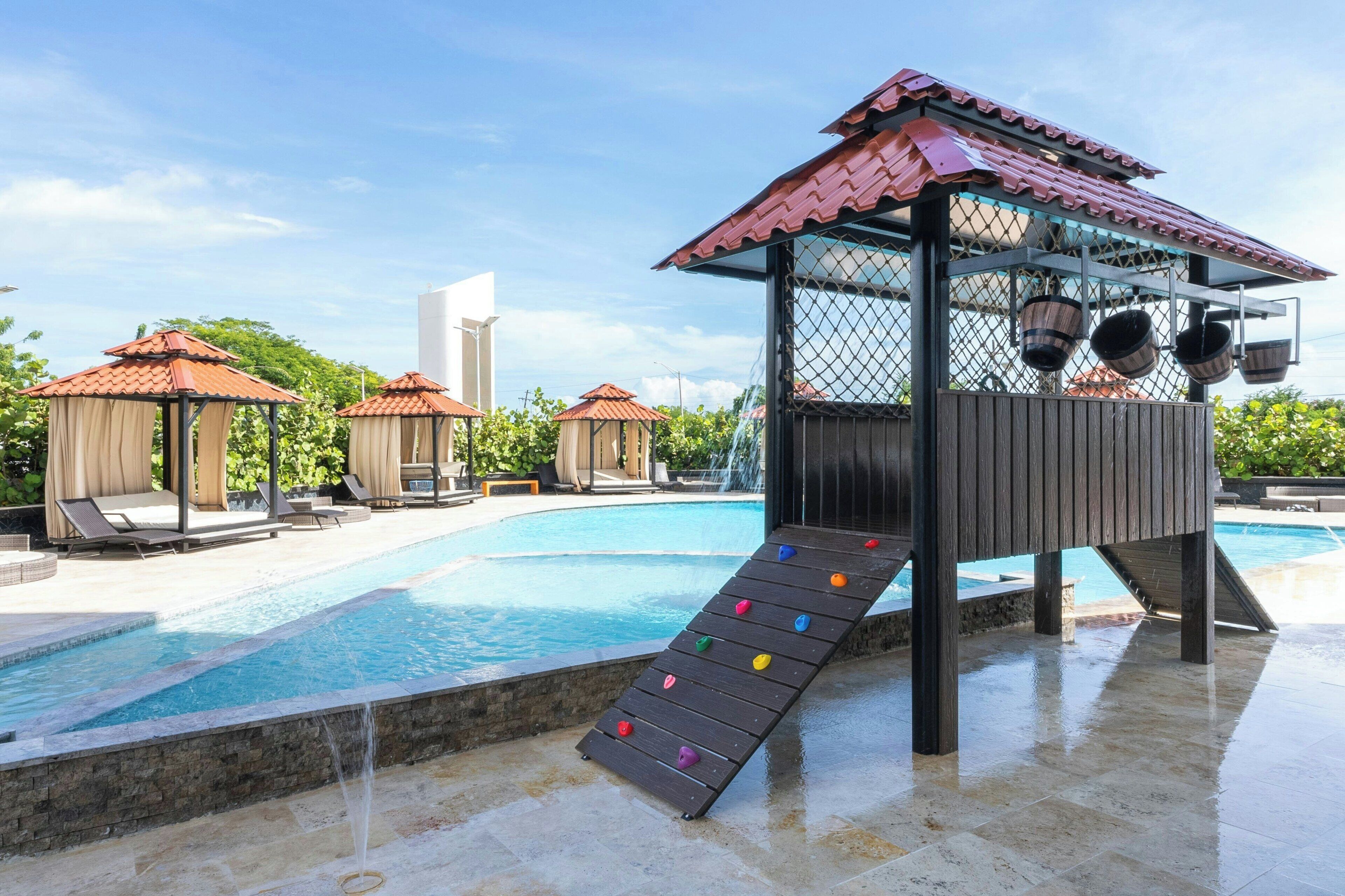 Piscina com escorregador de madeira e dois quiosques com telhados vermelhos.