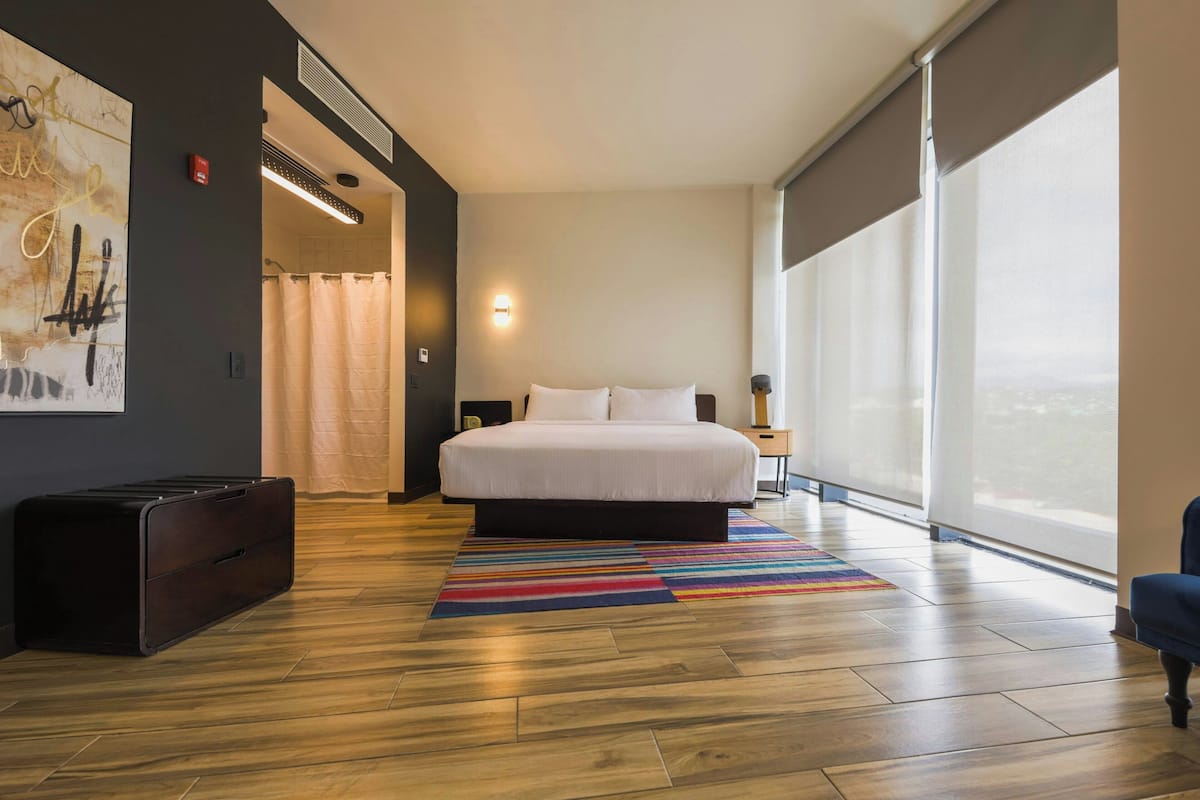 A modern hotel room with a large bed, a view through floor-to-ceiling windows, a colorful striped rug, and a dark-colored suitcase on the floor.
