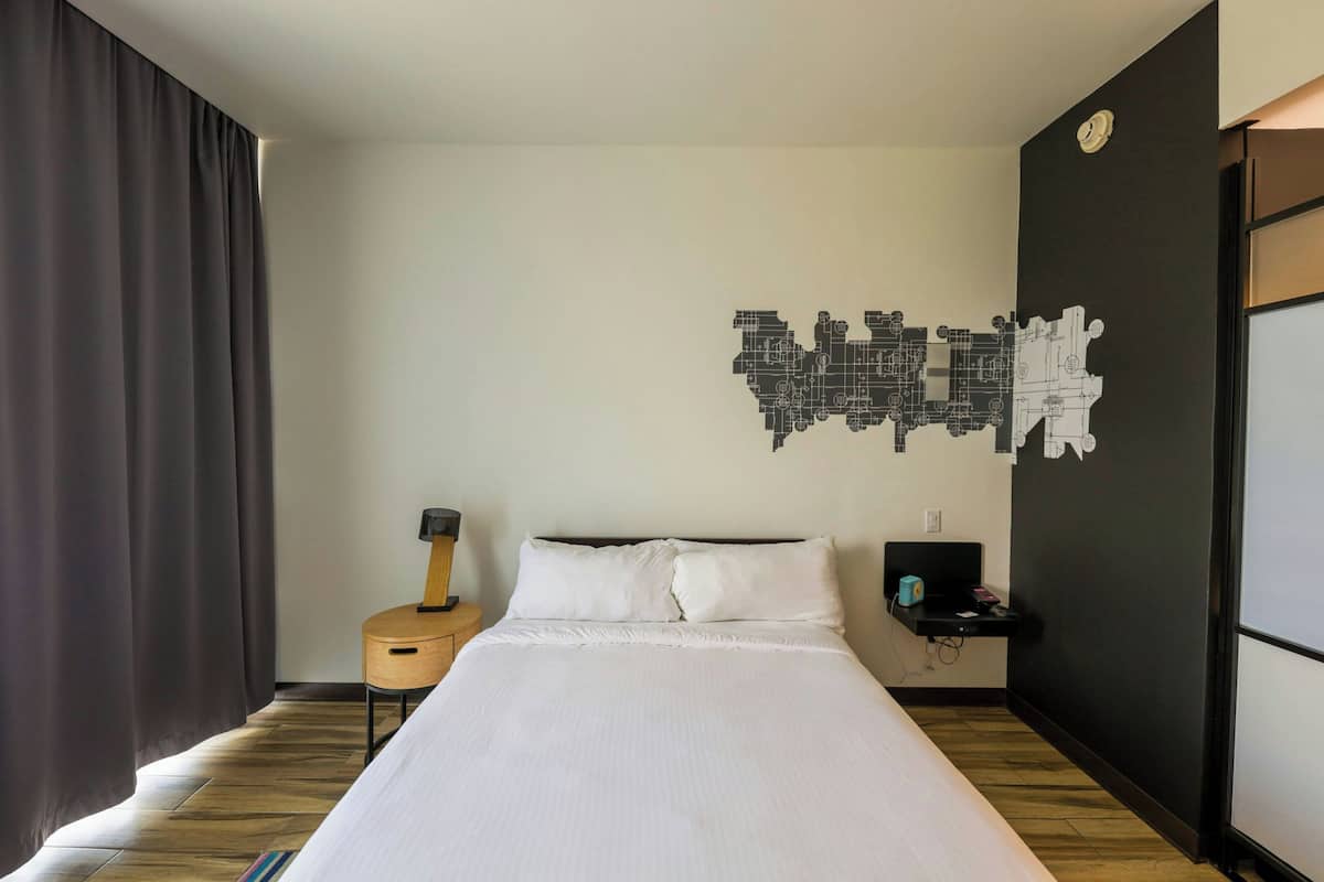 A hotel room with a large bed, a guitar, a small table with a lamp, and a wall art piece.