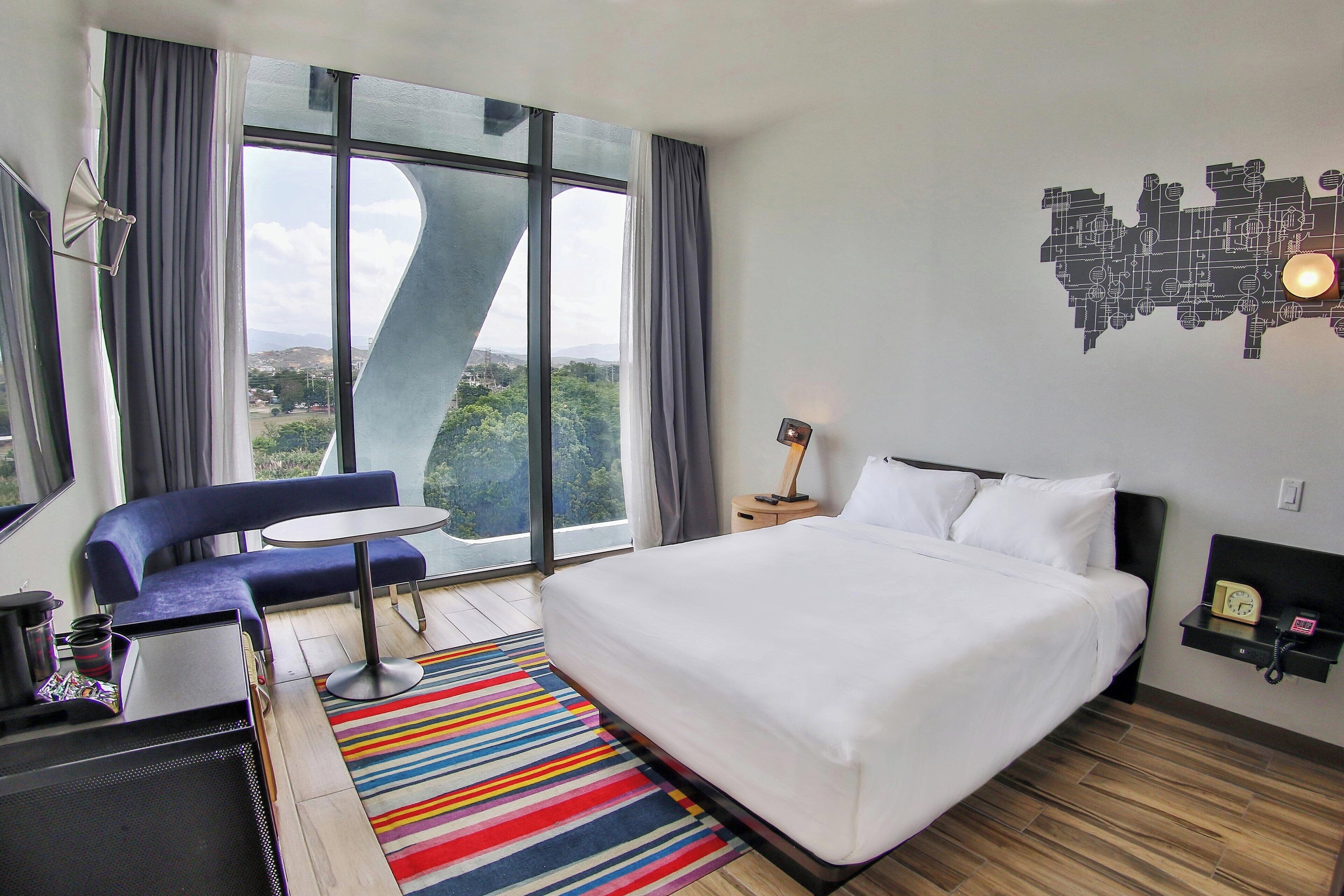 Una habitación de hotel moderna con una cama grande, un sofá azul, una mesita y vistas al exterior a través de amplios ventanales.