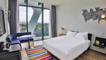 Una habitación de hotel moderna con una cama grande, un sofá azul, una mesita y vistas al exterior a través de amplios ventanales.