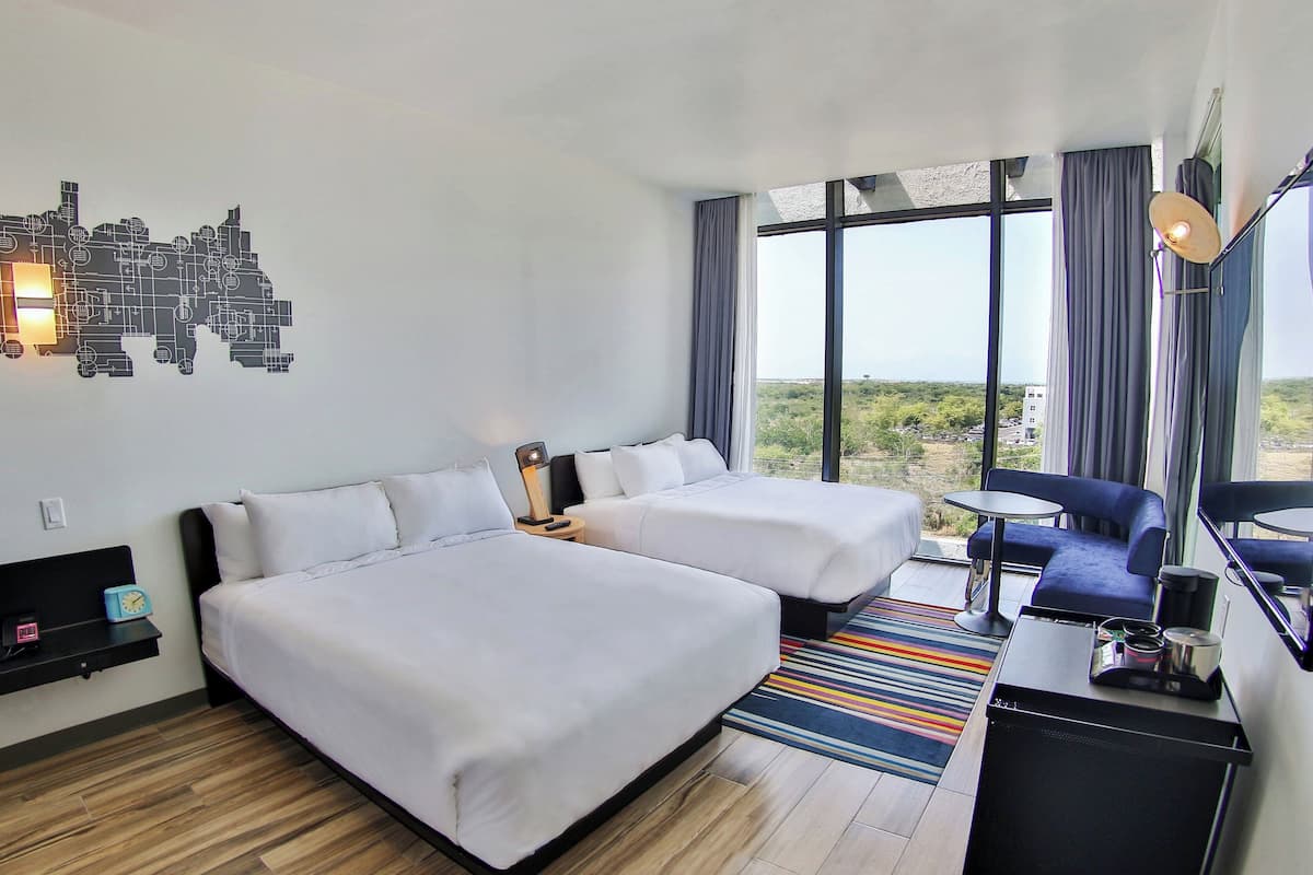 A hotel room with two beds, a small desk, a chair, and a large window offering a view of the outdoors.