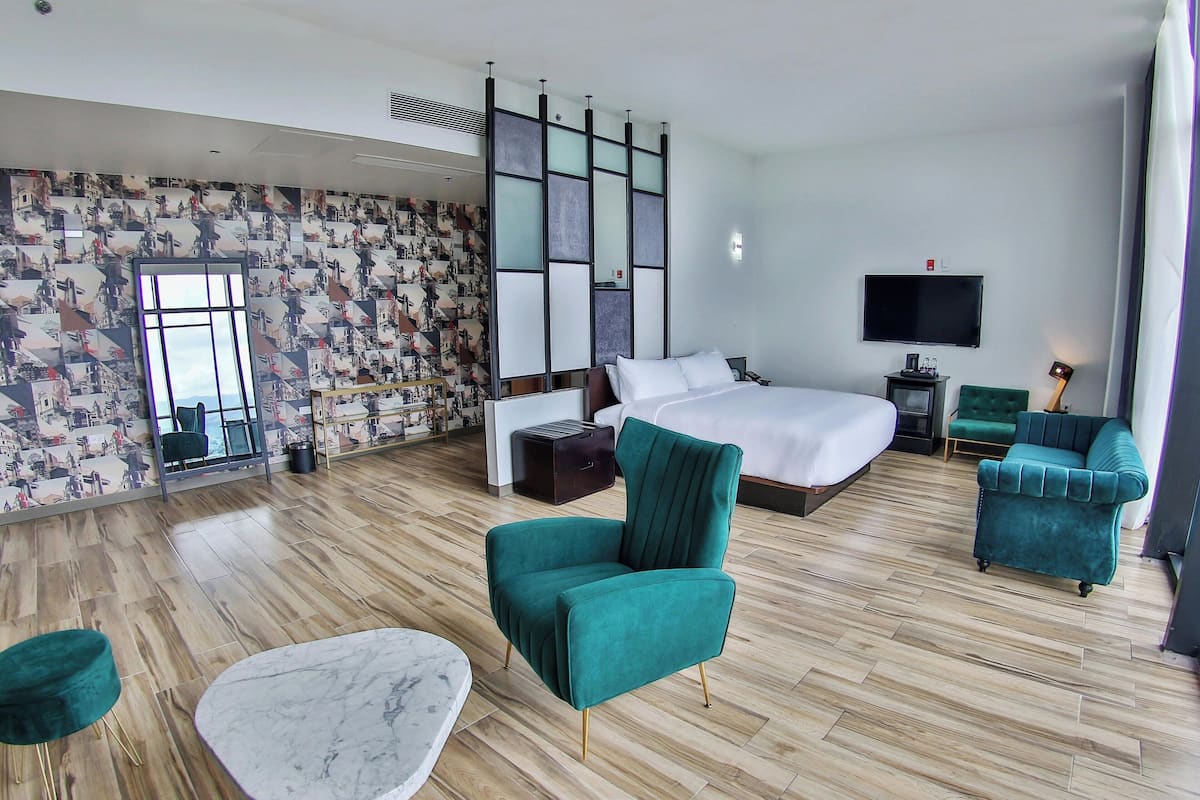 A modern hotel room with a large bed, a TV, a desk, and two green armchairs.