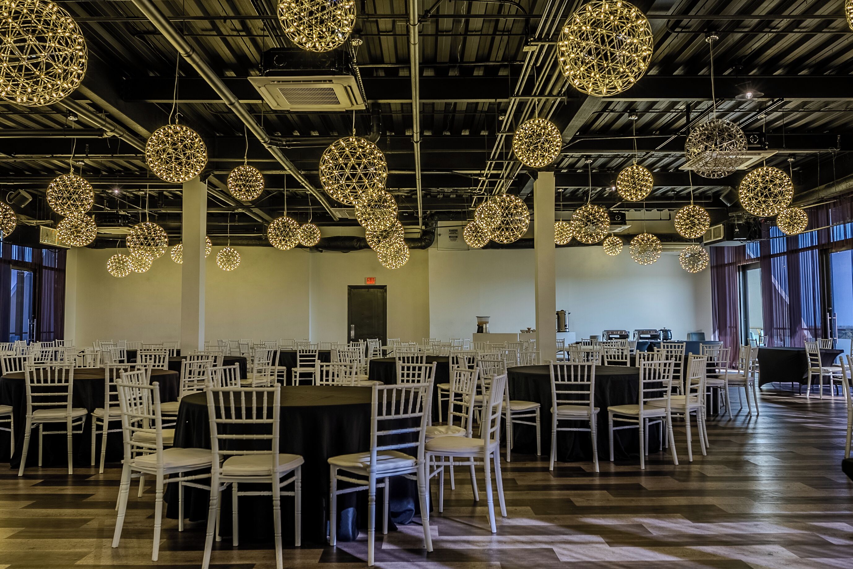 A spacious hall with round tables and chairs, large hanging spherical light fixtures, and a high ceiling with visible ductwork.