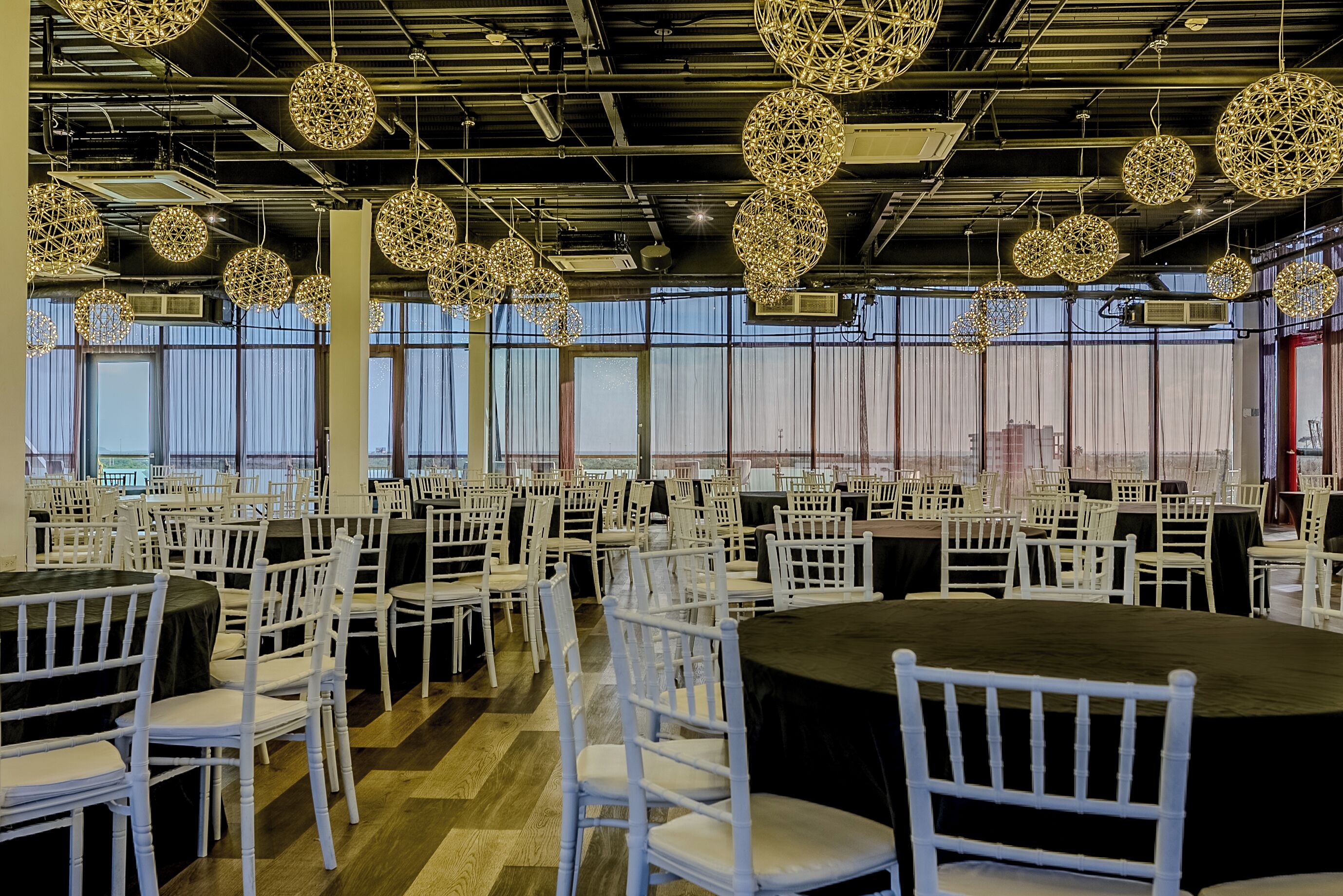 Un salón amplio con mesas redondas y sillas blancas, ventanas grandes y lámparas decorativas colgantes.