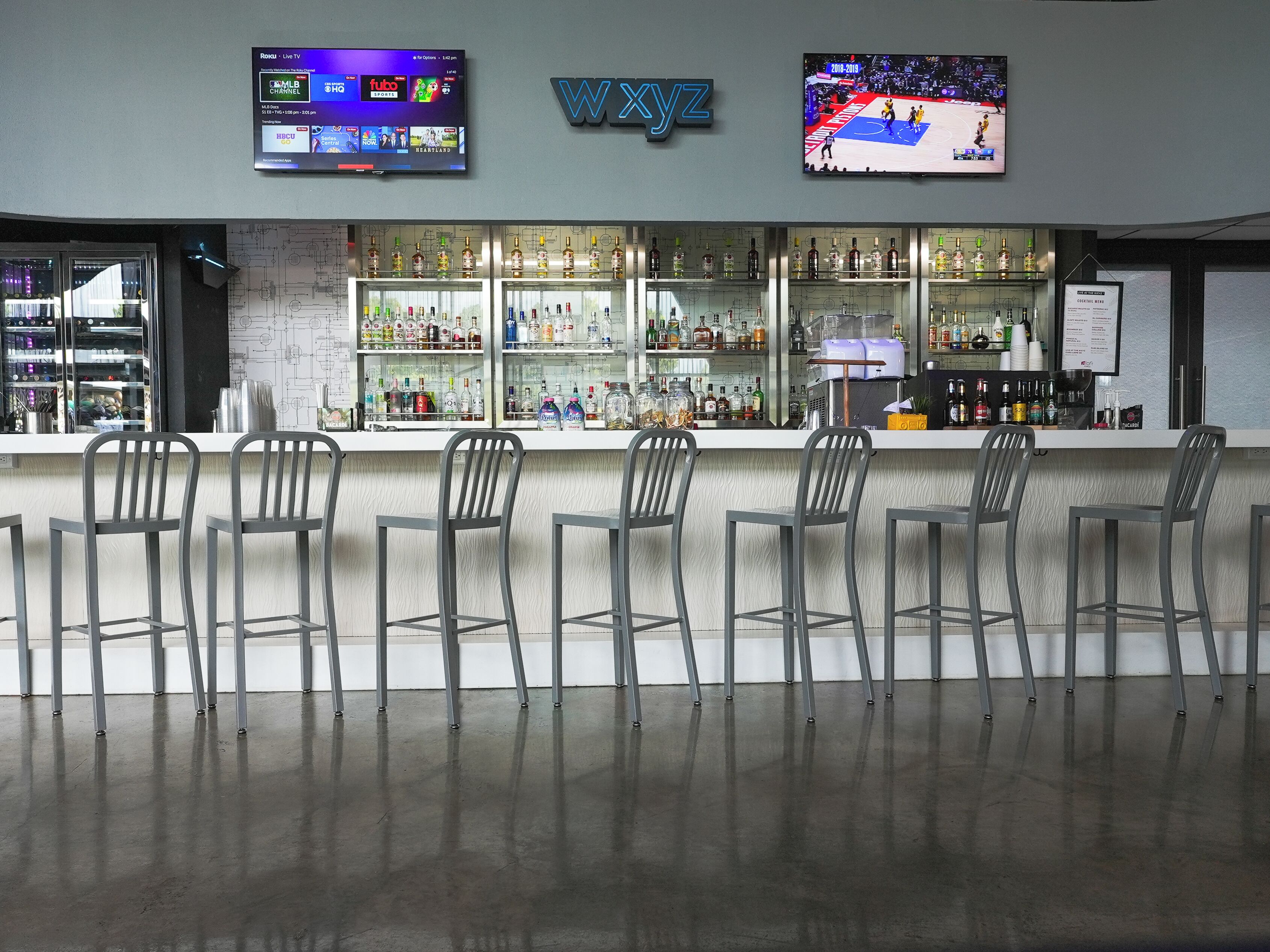 A bar area with a modern design, featuring a bar counter, multiple seating options, and a television mounted above the bar displaying sports content.